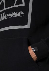 Czarna bluza z kapturem z szarym logo, z kieszenią z przodu i ściągaczami na rękawach. Materiał wydaje się miękki z delikatną fakturą.