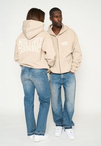 Sudaderas con cremallera en color beige con texto de logo blanco en la parte trasera, combinadas con jeans azules claros y zapatillas blancas. Diseño de outfit sencillo y casual.