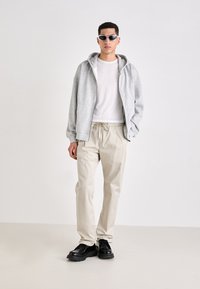 Grå zip-up hoodie over en hvid t-shirt, beige bukser med snøre og sorte snørebåndssko. Model iført mørke solbriller står imod en lys baggrund.