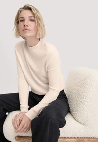 Cremfarbener Strickpullover mit hohem Kragen und gerippten Bündchen, getragen mit dunkelblauen, weiten Hosen und auf einem strukturierten, hellen Kissen sitzend.
