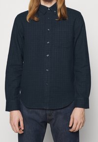 Tmavě modrá košile s dlouhým rukávem a knoflíkovým límcem, se jemným pruhovaným vzorem a jednou náprsní kapsou. Vyrobeno z texturované tkaniny.