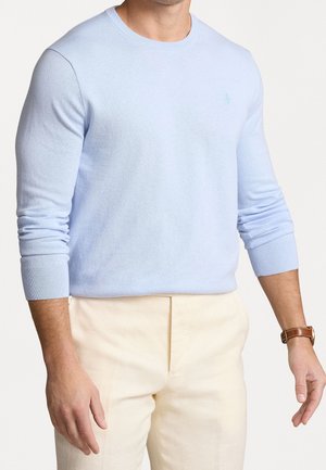 Mann trägt hellblauen Langarmpullover mit Rundhalsausschnitt und beige Hose, mit brauner Lederuhr am linken Handgelenk, steht mit entspannten Armen.