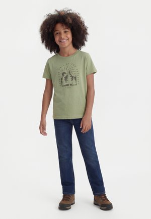 Uśmiechnięte dziecko z kręconymi włosami, ubrane w zielony t-shirt z grafiką, niebieskie dżinsy i brązowe buty trekkingowe, stojące na białym tle.