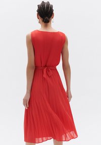Rode geplooide jurk met een mouwloos ontwerp, voorzien van een strikdetail in de taille en een vloeiende zoom. Gemaakt van een lichtgewicht stof.