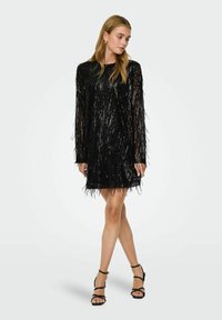 Melnas fringe kleita ar taisnu siluetu, garām piedurknēm un tekstūru detaļām. Izceļas ar īsu apakšējo malu un ir kombinēta ar melnajām sandālēm ar siksniņām.