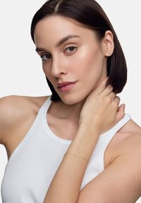 Złota bransoletka z małymi charmsami, noszona na nadgarstku osoby o jasnej karnacji. Osoba ma krótkie ciemne włosy i nosi biały top bez rękawów.