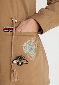 Tan jacket with a textured surface, featuring floral embroidery and a bee patch on the pocket, complemented by a braided drawstring.