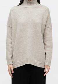 Femme portant un pull beige côtelé à col roulé avec manches longues sur un pantalon noir, debout devant un fond blanc uni.