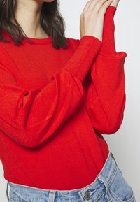 Roter Strickpullover mit langen Ärmeln, weichem Textil und figurbetonter Passform, kombiniert mit hellblauen, hochgeschnittenen Jeansshorts.