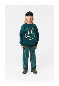 Felpa verde acqua con un design di faccina sorridente colorata, abbinata a pantaloni in corduroy verde acqua. Cappello di maglia nero e scarpe slip-on marroni.