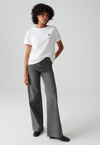 Weißes Baumwoll-T-Shirt mit kleinem schwarzem Herz-Logo, kombiniert mit grauen Jeans mit hoher Taille und weitem Bein. Schwarze Stiefeletten. Neutraler Hintergrund.