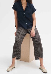 Kobieta siedząca na drewnianym bloku, ubrana w czarną, krótką koszulę zapinaną na guziki, wzorzyste szerokie spodnie oraz beżowe baleriny.