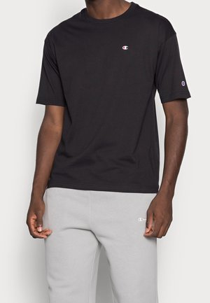 Mężczyzna w czarnym t-shircie z krótkim rękawem i jasnoszarych dresach, obie części z małymi logo marki Champion na klatce piersiowej, rękawie i udzie.