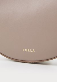 Okrugla, glatka torba od umjetne kože u prigušenoj mauve boji s zlatnim "FURLA" logotipom na dnu. Suptilna tekstura, minimalistički dizajn.