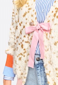 Cremefarbener Cardigan mit braunen Sprenkeln, der ein rosa Schleifchen-Detail und einen verzierten Akzent aufweist. Über einem blau gestreiften Hemd getragen.