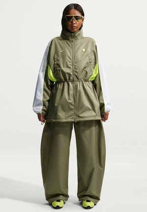 Kvinde iført oversized olivengrønt og hvidt træningssæt med neon gule detaljer og matchende sneakers, stående mod en ensfarvet baggrund.