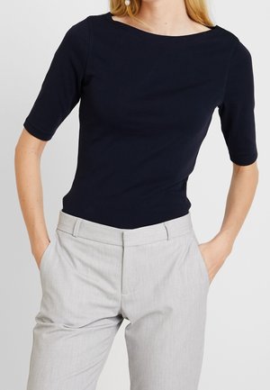 Kobieta ubrana w czarną bluzkę z krótkim rękawem i dekoltem w łódkę oraz jasno-szare, dopasowane spodnie, z rękami w kieszeniach, na jasnym, jednolitym tle.