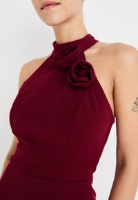 Burgundy mekko, jossa on korkea kaula-aukko, etuosassa kaksi kangaskukkaa. Valmistettu sileästä materiaalista, jossa on rakenteelliset saumat.