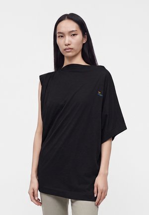 Femme aux longs cheveux noirs portant un haut noir asymétrique avec un petit logo coloré sur la poitrine, debout devant un fond uni.