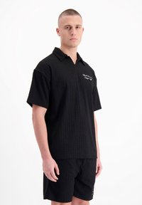 Zwarte getextureerde poloshirt met korte mouwen en een kraag, voorzien van een geborduurd logo op de borst, gecombineerd met bijpassende shorts in een vergelijkbaar materiaal.