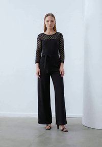 Mono negro con mangas largas de encaje y diseño de pata de elefante. Presenta un corsé ajustado, un cinturón de lazo en la cintura y una textura de tela suave.