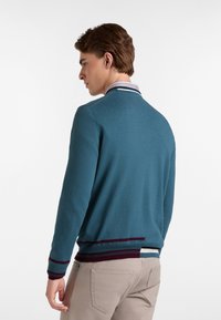 Cardigan lavorato a maglia turchese con colletto a righe e polsini accentuati in bordeaux, caratterizzato da una vestibilità comoda e una morbida consistenza.