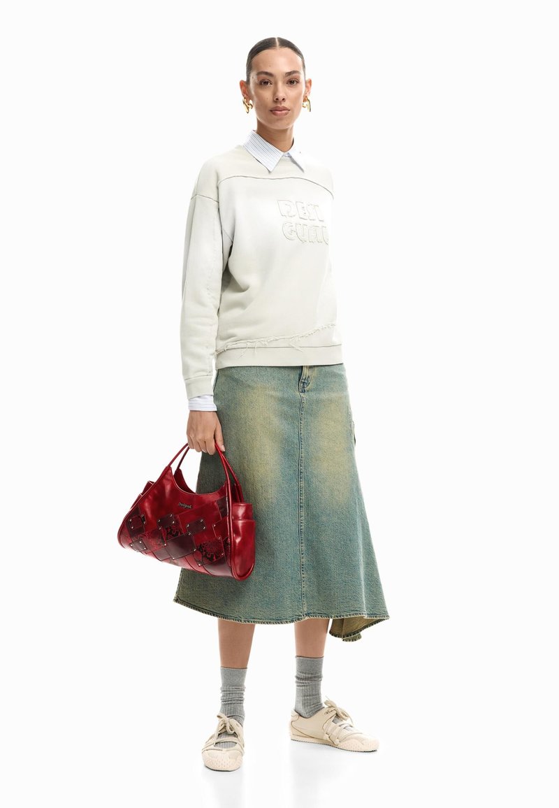 Fehér pulóvert viselő nő galléros inggel, farmer midi szoknyában, szürke zoknival, fehér tornacipővel, piros kézitáskát tartva, fehér háttér előtt áll.