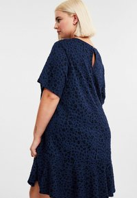 Blonde Frau in einem lockeren, strukturierten navyblauen Kleid mit schwarzen Leopardenflecken, die seitlich steht und eine Hand auf dem Saum des Kleides ruhen lässt.