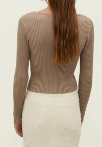Žena s dlouhými hnědými vlasy má na sobě těsný ribbed svetr v taupe barvě a sukně s vysokým pasem v odstínu off-white, je zobrazena zezadu.