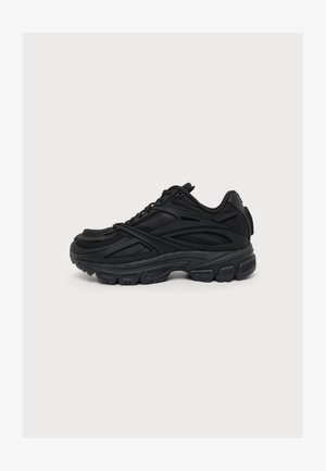 Zapatilla deportiva negra con suela texturizada, diseño con cordones y parte superior sintética en capas, mostrada de perfil sobre un fondo liso.