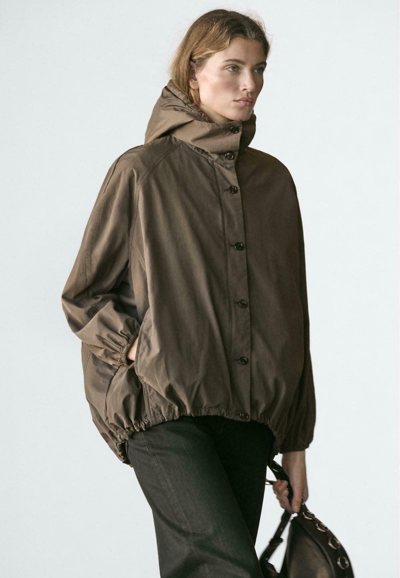 Femme portant une veste ample à capuche marron avec des boutons, un pantalon noir, tenant un sac à main noir devant un fond clair uni.