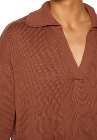 Rustrød strikket pullover med V-hals og kort krave. Teksturen er ribbet med en løs, afslappet pasform. Har ikke synlige metaldele.