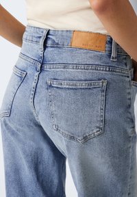 Ljust blå denimjeans med avslappnad passform, med bakfickor, synlig sömnad och en brun läderlogg på midjebandet.