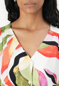 Short-sleeve blouse featuring a vibrant floral pattern in red, green, pink, and black, with a V-neck and white buttons.