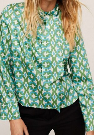 Frau trägt eine langärmelige Bluse mit grün-blauem geometrischem Hexagonmuster und großem Halstuchdetail, kombiniert mit schwarzer Hose.