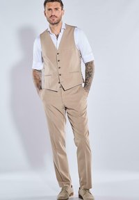 Beiger Anzug mit einreihiger Weste und Knopfdetails, kombiniert mit weißem Hemd und hellen Schuhen. Glatter Stoff und maßgeschneiderte Passform.