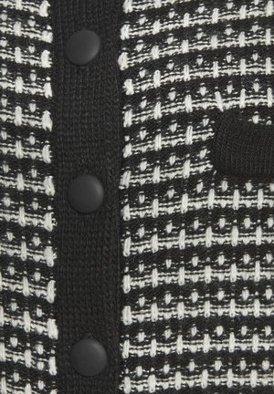Schwarz-weißer strukturierter Cardigan mit einem gewebten Muster, fünf schwarzen Knöpfen und einem gestrickten schwarzen Patch auf der Vorderseite.