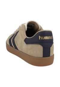 Beige Wildlederschuh mit marineblauen Akzenten, dicken Schnürsenkeln, Gummisohle und einem goldenen Logo an der Ferse. Die Seite zeigt ein Zickzackdesign.