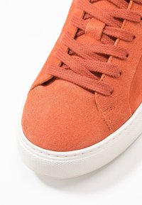 Oranžová semišová teniska s texturovaným povrchem, s plochými tkaničkami a bílou gumovou podešví. Detail špičky a oblasti tkaniček.
