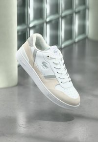 Λευκό και μπεζ αθλητικό παπούτσι Lacoste με κορδόνια, διάτρητο μύτη και λογότυπο στο πλάι, αιωρείται πάνω από τσιμεντένιο δάπεδο κοντά σε τοίχο από γυάλινα τούβλα.
