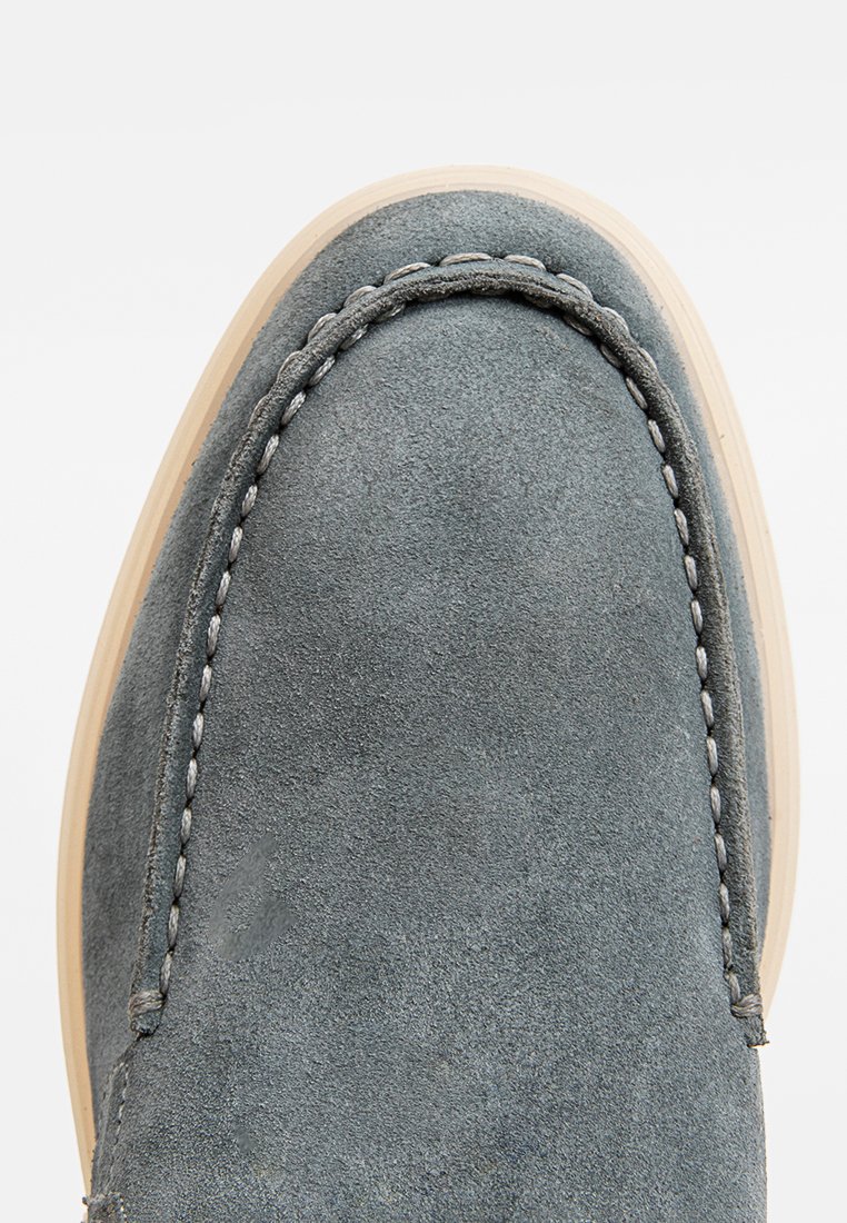 Chaussure en daim gris avec un bout arrondi, surpiqûres blanches contrastantes et semelle en caoutchouc de couleur claire. Texture lisse sans motifs.