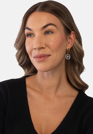 Frau mit hellbraunem, welligem Haar, trägt goldene und schwarze runde Ohrringe und ein schwarzes V-Ausschnitt-Top, leicht nach rechts blickend vor weißem Hintergrund.