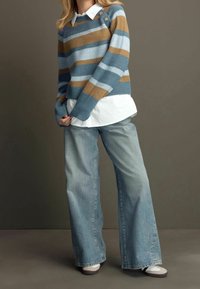 Pull en tricot rayé bleu et beige avec des poignets côtelés, porté par-dessus une chemise à col blanc, associé à un jean à jambes larges bleu clair et des baskets.