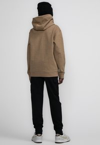 Hoodie marrón con diseño de logo grabado tono sobre tono, combinado con pantalones cargo negros. Terminando con un gorro negro y zapatillas blancas.