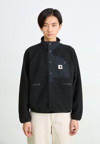 Persona con pelo corto negro, vistiendo una chaqueta de forro polar negra con cremalleras y bolsillos con botones, combinada con pantalones de color claro, de pie contra un fondo blanco.