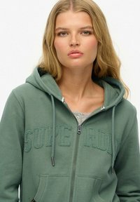 Groene rits hoodie met koordjes, met "SUPERDRY" geborduurd in een donkerdere tint. Zachte stof, voorzakken en geribbelde manchetten.
