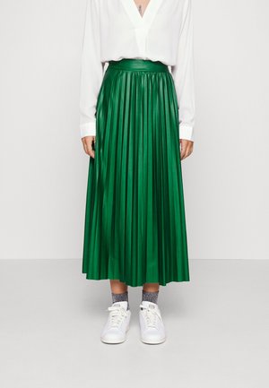 Frau trägt eine weiße langärmlige Bluse, grünen plissierten Midirock, graue Socken und weiße Sneaker, steht vor einem einfarbigen Hintergrund.