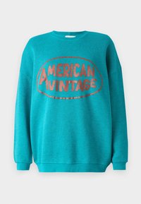 Turkos crewneck-tröja med långa ärmar och "AMERICAN VINTAGE" tryckt i stora, rundade röda bokstäver inuti en oval form.