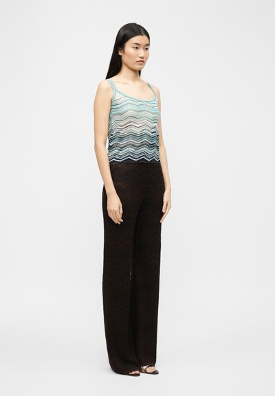 Γυναίκα που στέκεται, φορώντας μπλε τοπ χωρίς μανίκια με σχέδιο ζιγκ-ζαγκ και φαρδιά μαύρη παντελόνια, στραμμένη ελαφρώς προς τα δεξιά σε ουδέτερο φόντο.