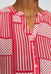 Chemise à motifs rouges et blancs avec un col à encoches, fermeture à boutons et divers designs géométriques, présentant des coutures et textures visibles.